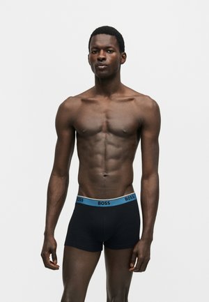 Atletische man staand, draagt zwarte boxer shorts met een blauwe tailleband met het label "BOSS", tegen een effen witte achtergrond.