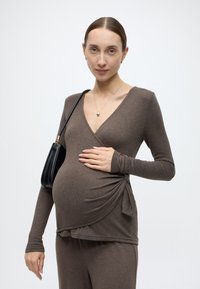 Καφέ μακρυμάνικη μπλούζα σε στυλ περιτύλιξης από απαλό ύφασμα με λεπτομέρεια ζέστας στη μέση, φορεμένη με παντελόνι που ταιριάζει. Μαύρη τσάντα χειρός και απλά κοσμήματα.