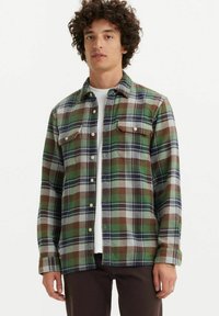 Camisa de franela a cuadros verde y marrón con dos bolsillos en el pecho, cierre frontal de botones y mangas largas. Presenta una textura suave y un corte casual.
