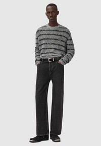 Man wearing a thick gray and black striped sweater, black wide-leg jeans with a belt, and black boots, standing with hands in pockets.