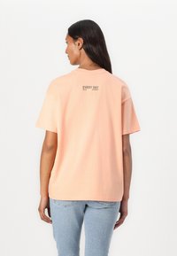 Camiseta de manga corta en algodón color durazno claro, de corte oversize, con texto en la parte posterior que dice "CADA DÍA ES UN INICIO" en verde apagado.