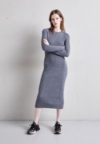 Una mujer se encuentra de pie con los brazos cruzados, vistiendo un largo vestido de punto gris acanalado y zapatillas negras, contra una pared lisa de color gris claro.