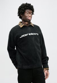 Μαύρο φλις πουλόβερ με λογότυπο "Jordan" σε λευκό, με υφή επιφάνειας, μακριά μανίκια και πουκαμισένιο γιακά με καρό μοτίβο από κάτω.