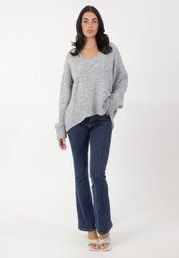 Grauer oversized Strickpullover mit V-Ausschnitt, geripptem Saum und weiten Ärmeln, kombiniert mit dunklen, ausgestellt geschnittenen Jeans und hellen, offenen Schuhen.