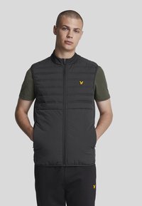 Gilet imbottito nero con cuciture verticali, colletto alto e zip frontale. Presenta un logo giallo sul lato sinistro del petto. Indossato sopra una camicia verde.