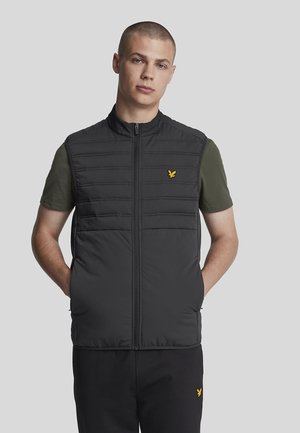 Gilet rembourré noir avec des coutures verticales, col haut et fermeture éclair avant. Présente un logo jaune sur la poitrine gauche. Porté par-dessus une chemise verte.