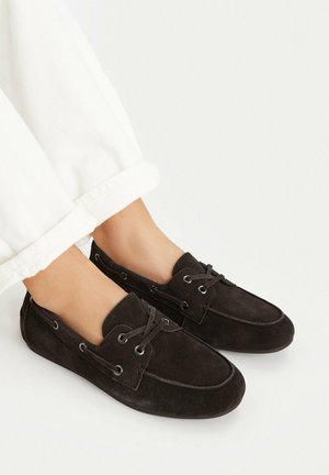 Stopy w czarnych zamszowych mokasynach z ozdobnymi sznurówkami, zestawione z białymi podwiniętymi spodniami na jednolitym tle.