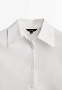 Chemise blanche à manches longues en tissu lisse, avec un col évasé classique et six boutons sur le devant. Étiquette visible.