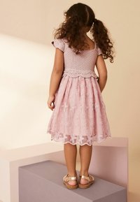 Helles rosa Kleid mit einem gehäkelten Oberteil, kurzen Ärmeln und einem mehrlagigen Tüllrock mit floralen Stickereien, kombiniert mit flachen goldenen Sandalen.