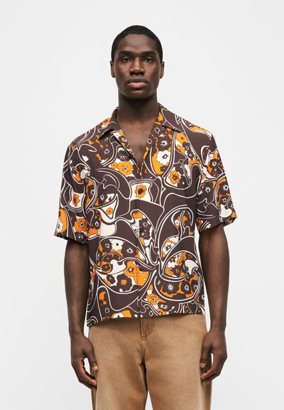 Άντρας όρθιος που φοράει καφέ, πορτοκαλί και λευκό πουκάμισο με κοντά μανίκια και λουλουδάτο μοτίβο, με ανοιχτό καφέ παντελόνι, μπροστά σε απλό φόντο.