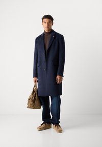 Abrigo de lana azul marino con cuello de muesca, cinco botones y dos bolsillos frontales, combinado con un suéter de cuello alto marrón y jeans azules. Zapatos beige y bolso marrón.