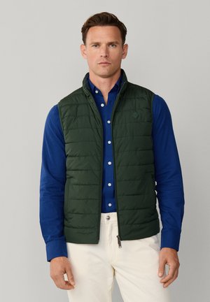 Mann trägt eine dunkelgrüne gesteppte Weste über einem blauen Hemd mit Knöpfen und hellen Hosen, steht vor einem schlichten Hintergrund.