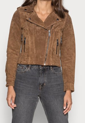 Femme portant une veste courte de biker en daim marron avec fermeture éclair légèrement décalée, associée à un jean noir délavé taille haute.