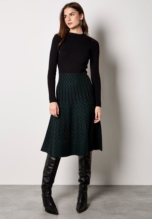 Vrouw die een zwarte geribbelde top met lange mouwen draagt, een groen patroon midi-rok en zwarte leren laarzen tot aan de knie, staand tegen een effen muur.