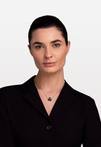 Zeltīta kaklarota ar melnu ziedu piekariņu, kas sastāv no trim ziedlapām. Modelis valkā melnu žaketi ar apkakli uz neitrāla fona.