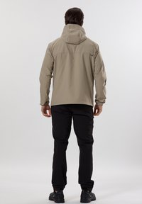 Veste étanche beige clair avec capuche ajustable, poignets élastiques et coupe ample. Associée à un pantalon noir et des chaussures sombres.