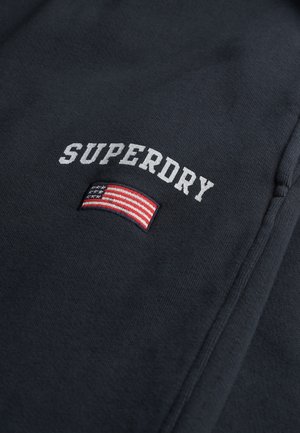 Sweatshirt bleu marine foncé avec "SUPERDRY" brodé en blanc. Comprend un écusson du drapeau américain avec des rayures rouges, blanches et bleues.
