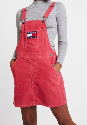 Woman wearing a pink corduroy overall dress with front pocket and "Tommy Jeans" logo, paired with a light gray ribbed long-sleeve top.