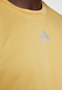 Detailní záběr na žlutou košili s jemně strukturovanou látkou a malým světle šedým trojúhelníkovým logem se třemi pruhy na horní části hrudníku.