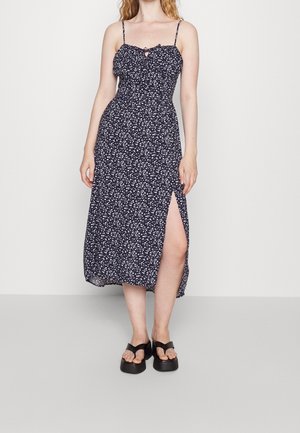 Kobieta w czarnej sukience midi w białe kwiatowe wzory, z cienkimi ramiączkami, rozcięciem z przodu i czarnymi platformami na sandałach, stojąca na jednolitym tle.