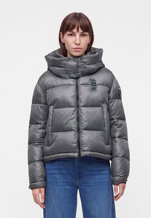 Frau trägt eine graue, gesteppte Jacke mit hohem, geknöpftem Kragen und blaue Jeans, steht vor einem einfarbigen hellen Hintergrund.