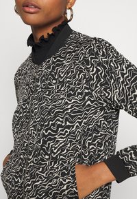 Bomber bunda v černé a krémové barvě s vlnkovým vzorem, vybavená zipovým zapínáním, žebrovanými manžetami a bočními kapsami. Texturovaná tkanina s hladkým povrchem.