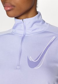 Vaaleanlila puolivetoketjupaita, joka on valmistettu pehmeästä kankaasta. Paita sisältää erottuvan valkoisen Nike-logon ja korkean kaulusmuotoilun.
