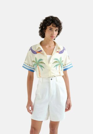 Mujer con cabello corto y rizado que lleva una camisa crema con estampados de palmera y nadador, y pantalones cortos blancos de cintura alta sobre un fondo liso.