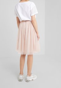 Mujer vestida con una blusa de manga corta blanca de puntillas, una falda midi de tul rosa claro y zapatillas blancas, de pie contra una pared lisa.