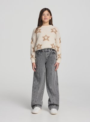 Maglione color crema con motivi a stella marroni, abbinato a jeans grigi a vita alta e gamba larga, e sneaker bianche. Texture complessivamente liscia.