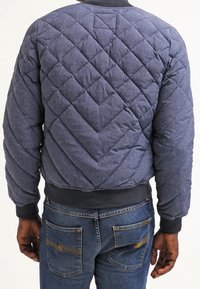Modrá prošívaná bomber bunda s diamantovým vzorem, žebrovanými manžetami a lemem, a uvolněným střihem. Nosená s tmavě modrými džíny.
