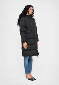 Black puffer coat, knee-length, with a zippered front and side pockets. Smooth texture, and a hood, styled with fitted blue jeans and black heels.