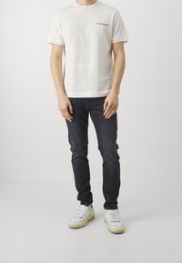 Biała bawełniana koszulka z krótkim rękawem i czerwonym logo na lewej piersi, zestawiona z ciemnymi dżinsami i wielokolorowymi sneakersami.