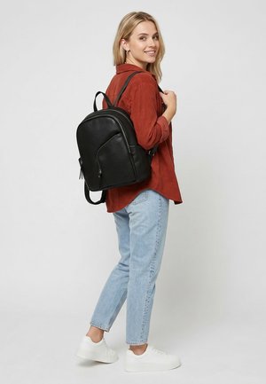 Smilende kvinde iført en rustrød skjorte, lyseblå jeans, hvide sneakers og bærende en sort læderrygsæk på ryggen.