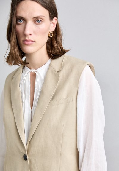 Gilet sans manches beige en tissu texturé, à un bouton et col cranté, associé à une blouse blanche à manches longues avec un col en lien.