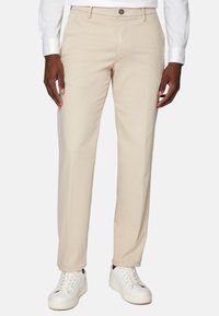 Bēgu bikses ar gludu auduma tekstūru, klasiskā taisnā piegriezumā un ar pogu aizdari, kombinētas ar baltām sporta apaviem.