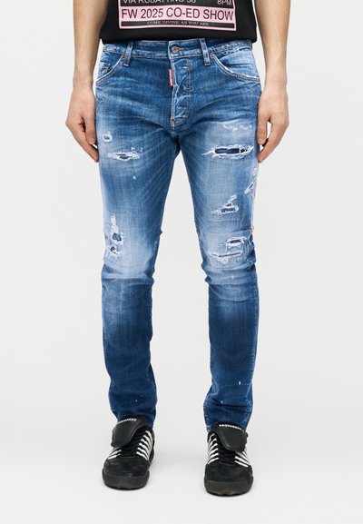 Άνδρας που φοράει μπλε στενά τζιν με φθορές και μαύρα αθλητικά παπούτσια με λευκά κορδόνια, στέκεται μπροστά σε ένα απλό λευκό φόντο.