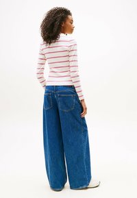 Femme aux cheveux bouclés portant un haut à manches longues blanc avec des rayures rouges et un jean bleu à large jambe, se tenant contre un fond blanc.