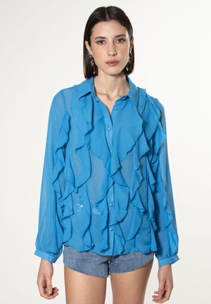 Blouse bleue, transparente, avec un design à volants, fermeture à boutons à l'avant et manches longues. Le matériau est léger ; elle dispose d'un col et d'une légère brillance.