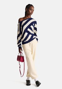 Maglione a righe navy e crema con design off-shoulder, abbinato a pantaloni a gamba larga crema e una piccola borsa rossa. Indossati mocassini neri.