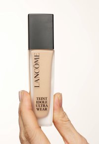 Flacon de fond de teint avec un bouchon noir rectangulaire et un contenu beige, étiqueté "Teint Idole Ultra Wear" en caractères gras noirs.
