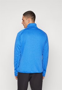 Blå fleece zip-up jakke med høy krage og lange ermer. Har et teksturert mønster og en rett kant. Brukt med sorte bukser.