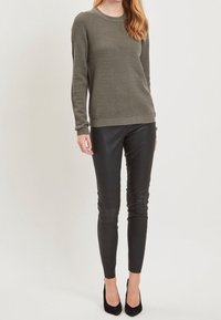 Olivgrön stickad tröja med ett strukturerat mönster, rund halsringning och matchande ribbade ärmslut, kombinerad med svarta åtsittande läderbyxor och svarta klackskor.