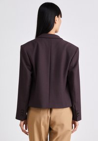 Blazer morado a medida con líneas de hombros marcadas, solapas con muesca y cuatro botones negros en las mangas, combinado con pantalones beige.