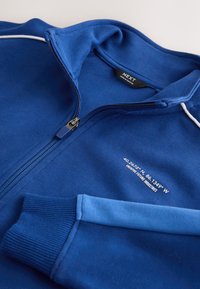 Veste zippée bleue avec poignets côtelés, passepoil blanc sur l'épaule, et texte blanc avec coordonnées et étiquette de marque à l'intérieur du col.