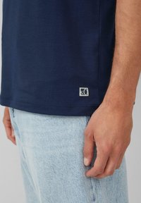 Granatowa koszulka z krótkim rękawem o gładkiej fakturze, z małym logo marki w kontrastowym odcieniu near do dołu, w zestawieniu z jasnymi dżinsami.