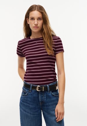 Jeune femme aux longs cheveux châtain clair portant un t-shirt violet rayé et un jean bleu avec une ceinture noire, debout sur un fond blanc.