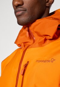 Mies, który ma na sobie jasnopomarańczową kurtkę z wysokim kołnierzem zapinanym na zamek oraz haftowanym logo Norrøna na piersi.