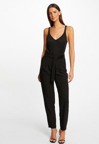 Bodysuit noir à décolleté en V, avec des accents argentés sur les bretelles, associé à un pantalon noir taille haute avec un détail en nœud à l'avant et des jambes fuselées.