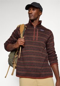 Red and green patterned fleece pullover with a zip neck, orange accents, and ribbed cuffs. Worn with a tan backpack.